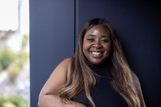 Image of a young black woman smiling at the camera. She is stood against a dark blue wall, and a blurred background.